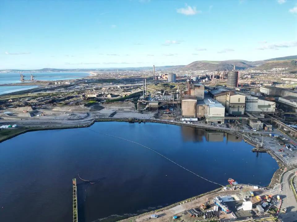 Aerial view of the Port Talbot steelworks, with a large water reservoir in the foreground, blast furnaces and chimneys beyond, and Swansea Bay in the distance.