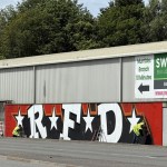 Giant graffiti letters RFD with white stars on a red and black background being painted on the front wall of St Helen's stadium, with two painters in hi-vis vests at work