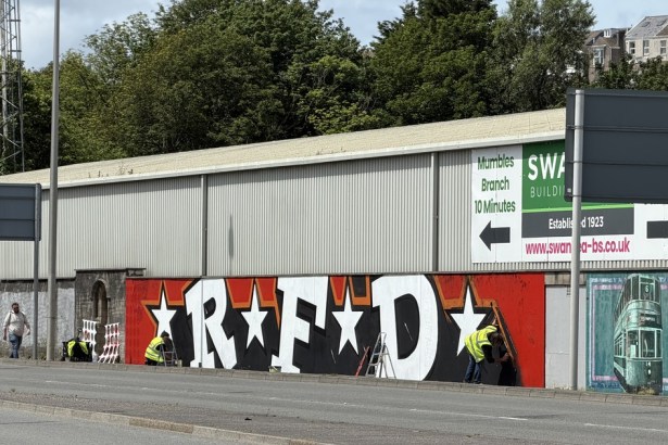 Giant graffiti letters RFD with white stars on a red and black background being painted on the front wall of St Helen's stadium, with two painters in hi-vis vests at work