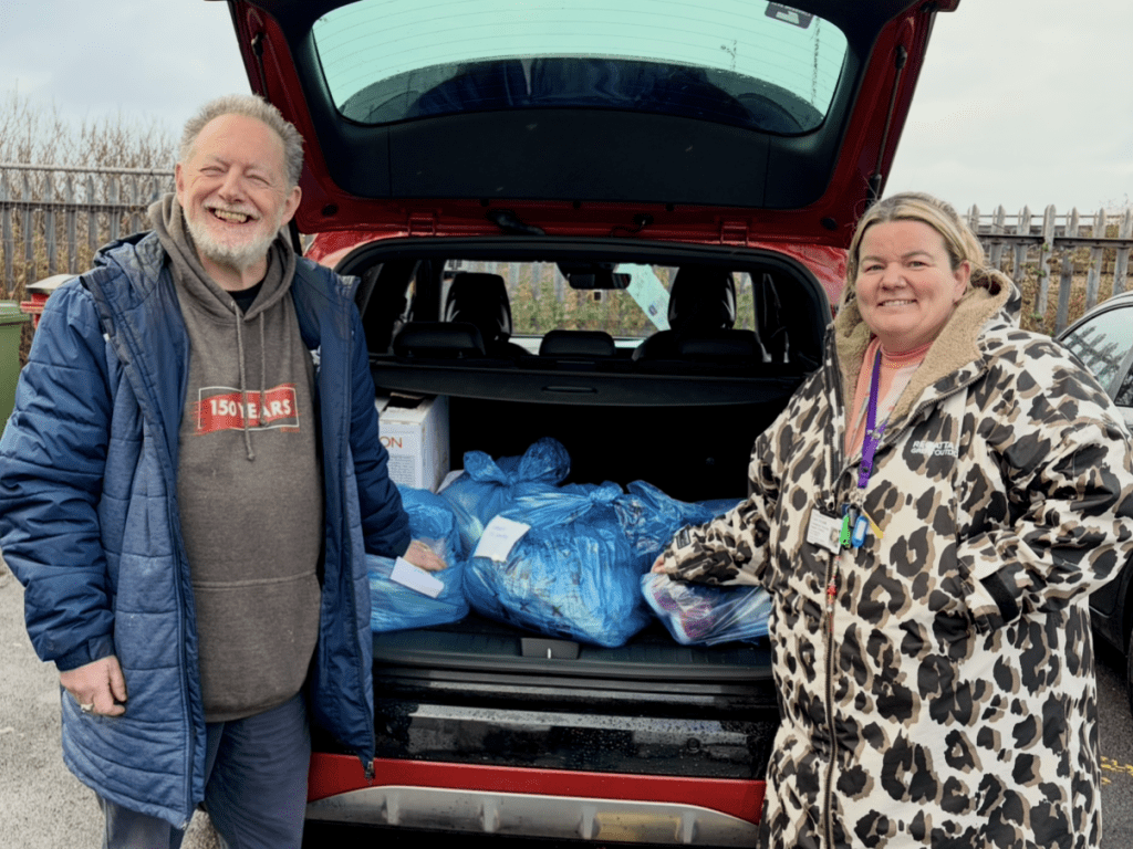 A man and a woman stand beside a car boot filled with blue bags of donated goods.