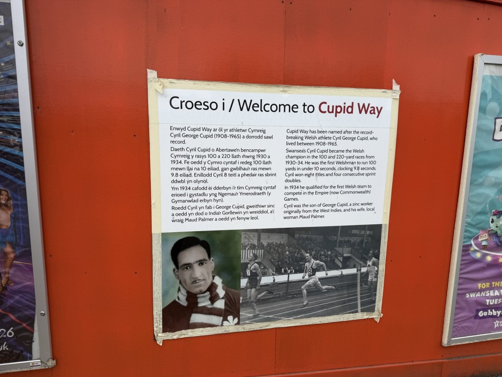 An information board headed "Welcome to Cupid Way" on red hoarding, with photos of 1930s Swansea sprinter Cyril Cupid.