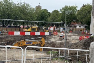 Excavation work in Castle Square behind site fencing, with a Thomas Plant Hire digger and the ruins of Swansea Castle behind.
