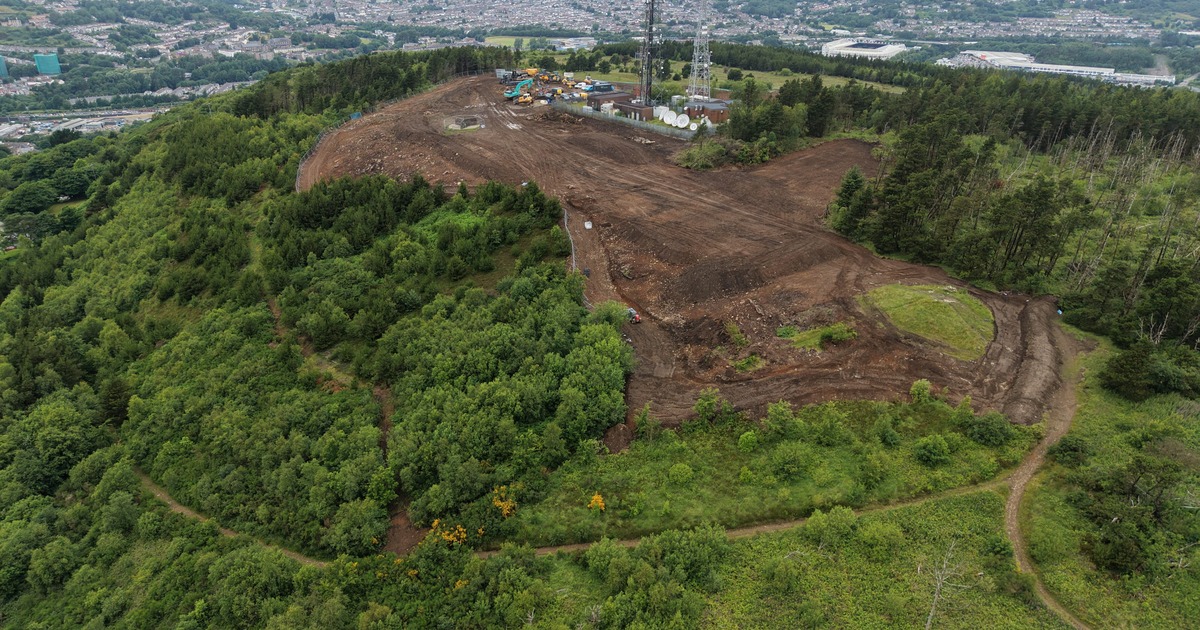 An aerial view of Kilvey Hill showing a large area of cleared ground and earthworks near the summit masts, with Swansea and the stadium in the distance.
