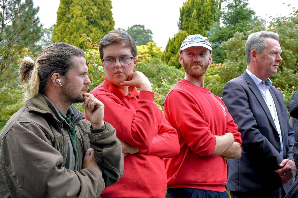 Three Swansea Council gardeners in red tops stand beside a man in a suit among trees and shrubs at the gardens.