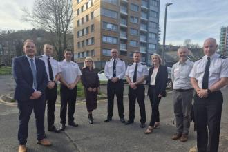 A group of eight people, including police officers in uniform and people in business dress, standing together outdoors in front of the Dyfatty flats.