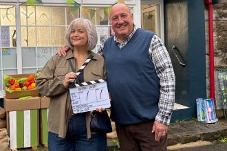 Ruth Jones and Steve Speirs smiling outside a village shop, Jones holding a clapperboard reading "Better Later", on the comedy's first day of filming.