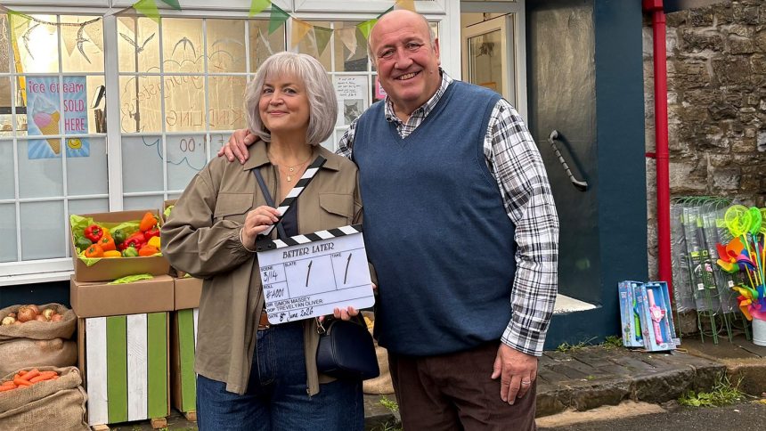 Ruth Jones and Steve Speirs smiling outside a village shop, Jones holding a clapperboard reading "Better Later", on the comedy's first day of filming.