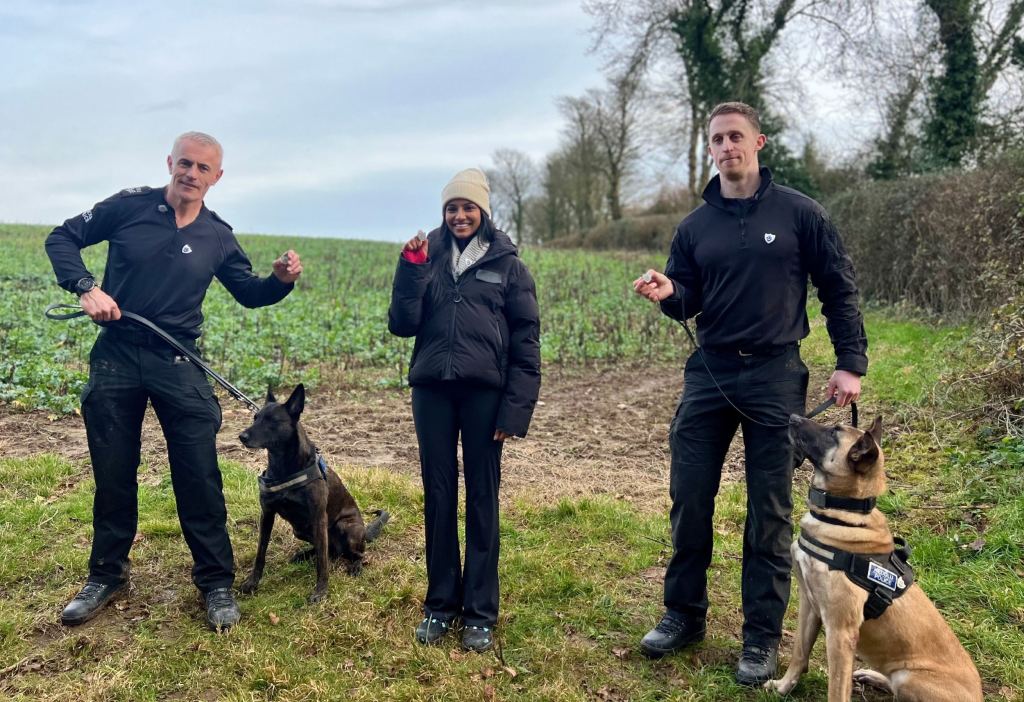 Two dog handlers and a presenter stand with two police dogs in a field.