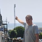 A man smiling and pointing up towards the tall steel St Helen's floodlight, with flowerbeds in the foreground and Swansea Bay behind