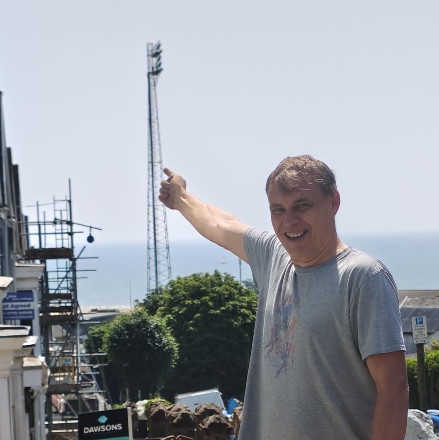 A man smiling and pointing up towards the tall steel St Helen's floodlight, with flowerbeds in the foreground and Swansea Bay behind