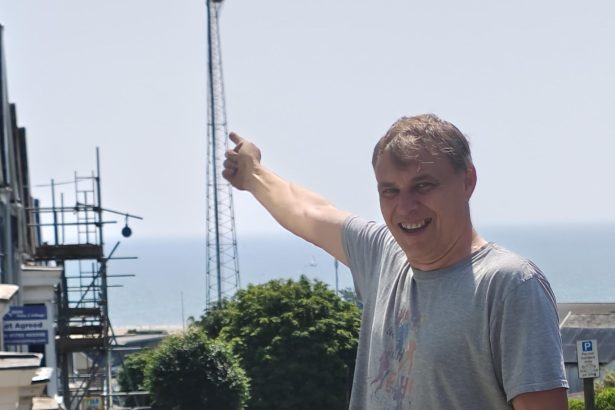 A man smiling and pointing up towards the tall steel St Helen's floodlight, with flowerbeds in the foreground and Swansea Bay behind