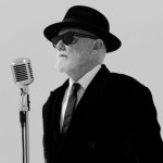 Roger Taylor in a black fedora, dark sunglasses, black velvet suit and tie, standing at a vintage silver microphone against a plain background, in black and white