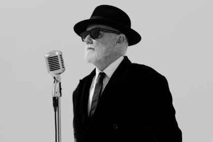 Roger Taylor in a black fedora, dark sunglasses, black velvet suit and tie, standing at a vintage silver microphone against a plain background, in black and white