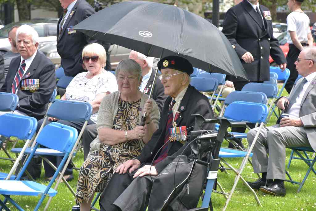 An elderly veteran in a beret and medals sits beneath an umbrella beside a woman, among rows of seated guests on the Guildhall lawn.