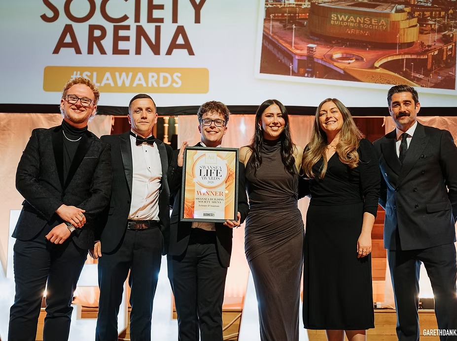 Five members of the Swansea Building Society Arena team holding a framed winner's certificate on stage at the Swansea Life Awards, alongside a presenter.