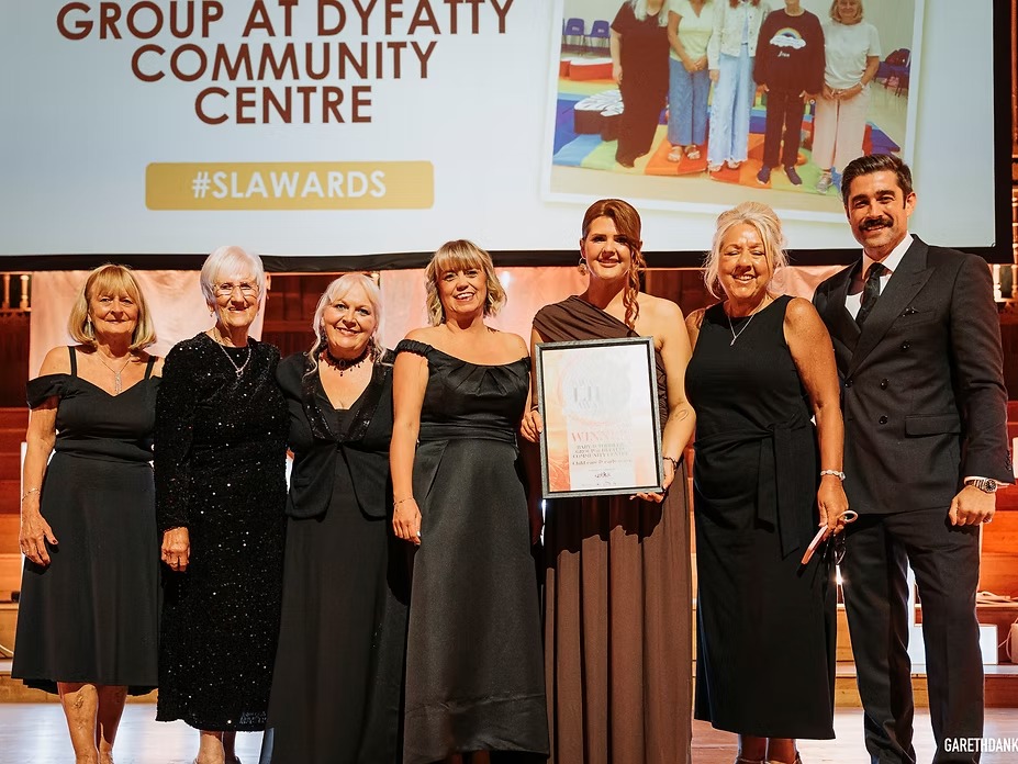 Six women holding a framed winner's certificate on stage at the Swansea Life Awards, with a presenter in a suit, in front of a screen reading "group at Dyfatty Community Centre".
