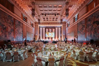 Round banqueting tables set for the Swansea Life Awards dinner in the Brangwyn Hall, with the Brangwyn Panels on the walls and a stage with "Swansea Life" lit letters.