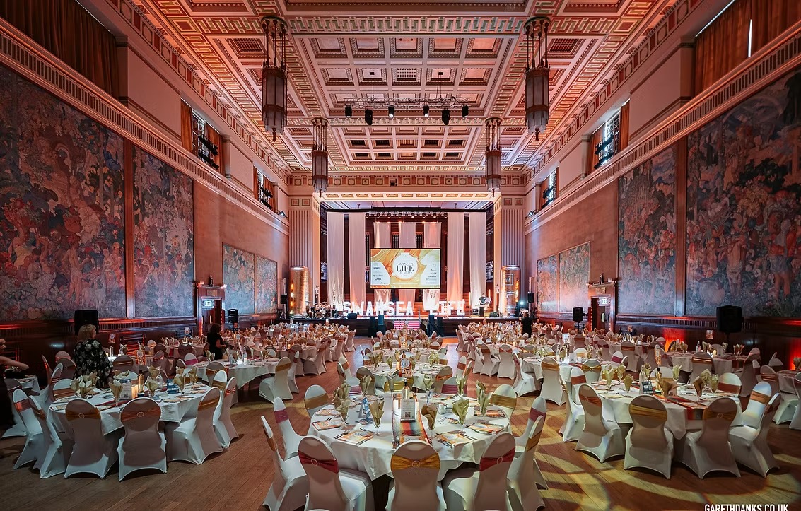 Round banqueting tables set for the Swansea Life Awards dinner in the Brangwyn Hall, with the Brangwyn Panels on the walls and a stage with "Swansea Life" lit letters.