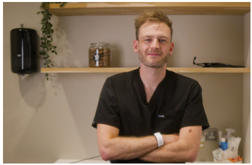 A vet in black scrubs stands with his arms folded in a clinic.