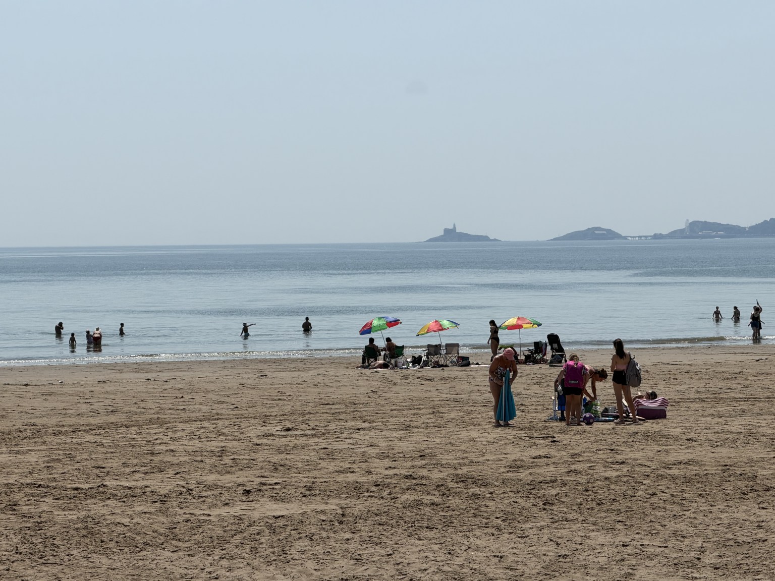 Crowds on Swansea beach during the June 2026 heatwave, with people swimming, parasols up and Mumbles lighthouse visible across the bay.