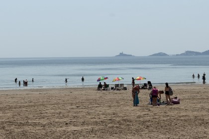 Crowds on Swansea beach during the June 2026 heatwave, with people swimming, parasols up and Mumbles lighthouse visible across the bay.