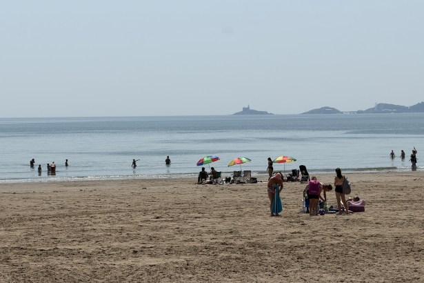 Crowds on Swansea beach during the June 2026 heatwave, with people swimming, parasols up and Mumbles lighthouse visible across the bay.