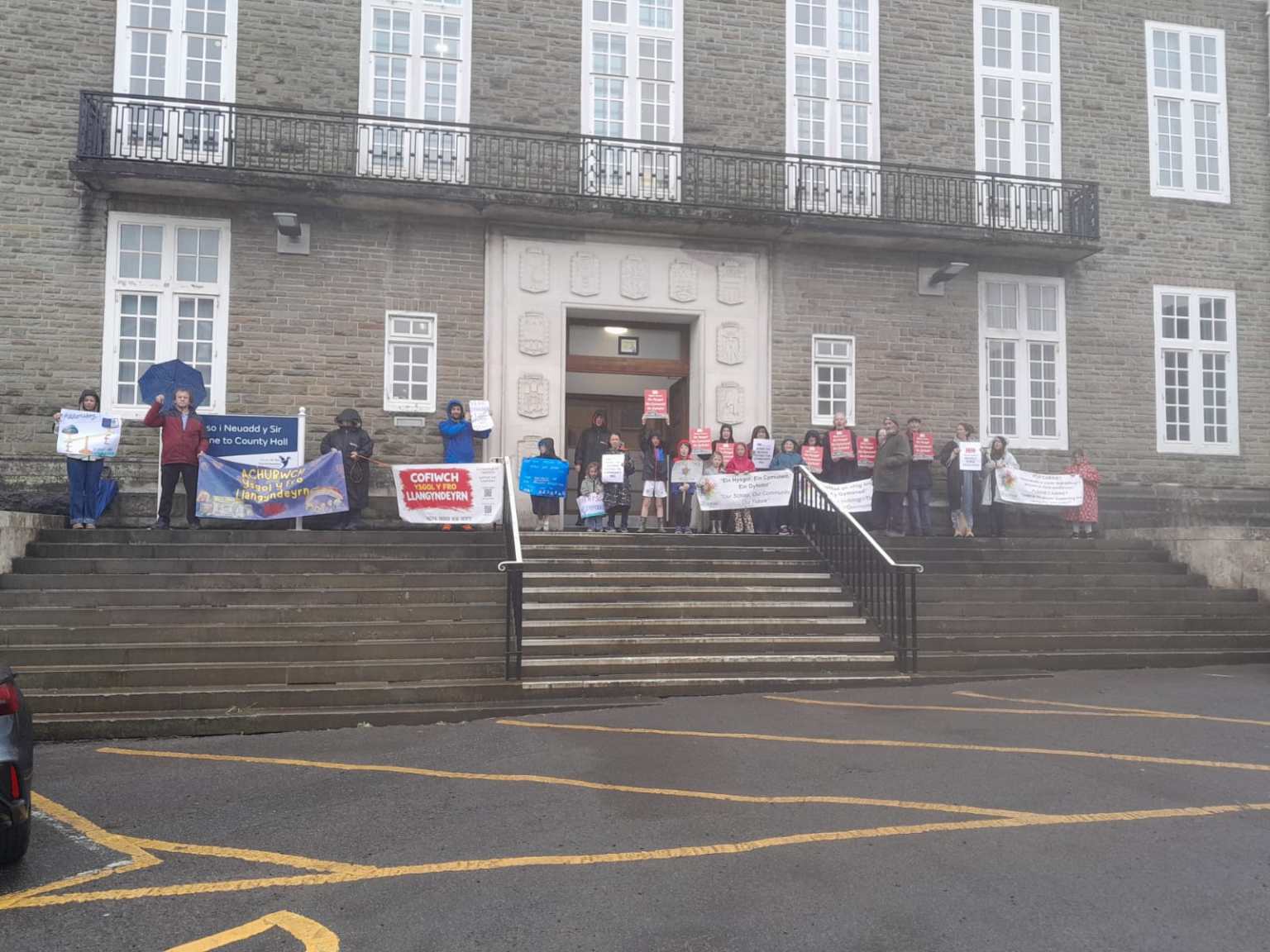 A group of protesters, including children, stand on the steps of County Hall in Carmarthen holding banners and placards opposing the closure of Ysgol y Fro, Llangyndeyrn, on a wet day.