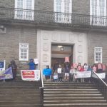 A group of protesters, including children, stand on the steps of County Hall in Carmarthen holding banners and placards opposing the closure of Ysgol y Fro, Llangyndeyrn, on a wet day.