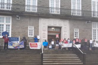 A group of protesters, including children, stand on the steps of County Hall in Carmarthen holding banners and placards opposing the closure of Ysgol y Fro, Llangyndeyrn, on a wet day.