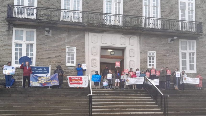 A group of protesters, including children, stand on the steps of County Hall in Carmarthen holding banners and placards opposing the closure of Ysgol y Fro, Llangyndeyrn, on a wet day.