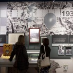 Two visitors look at wall displays at the Dylan Thomas Centre, including a large 1939 panel with aerial photographs, a vintage radio and framed documents.