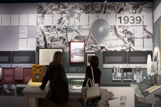 Two visitors look at wall displays at the Dylan Thomas Centre, including a large 1939 panel with aerial photographs, a vintage radio and framed documents.