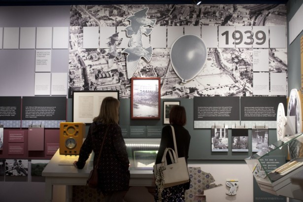 Two visitors look at wall displays at the Dylan Thomas Centre, including a large 1939 panel with aerial photographs, a vintage radio and framed documents.