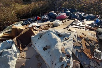 A large pile of fly-tipped waste including a mattress, cardboard, plasterboard and black bin bags on overgrown land.