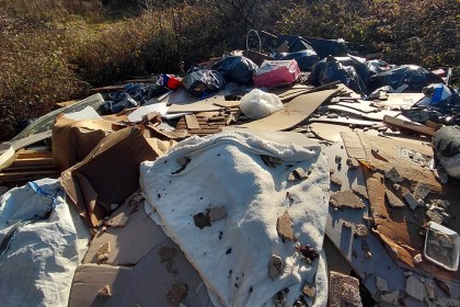 A large pile of fly-tipped waste including a mattress, cardboard, plasterboard and black bin bags on overgrown land.
