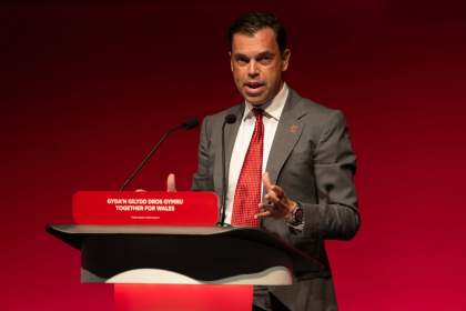 Ken Skates in a grey suit and red tie speaking at a red Welsh Labour lectern reading Gyda'n Gilydd Dros Gymru — Together for Wales