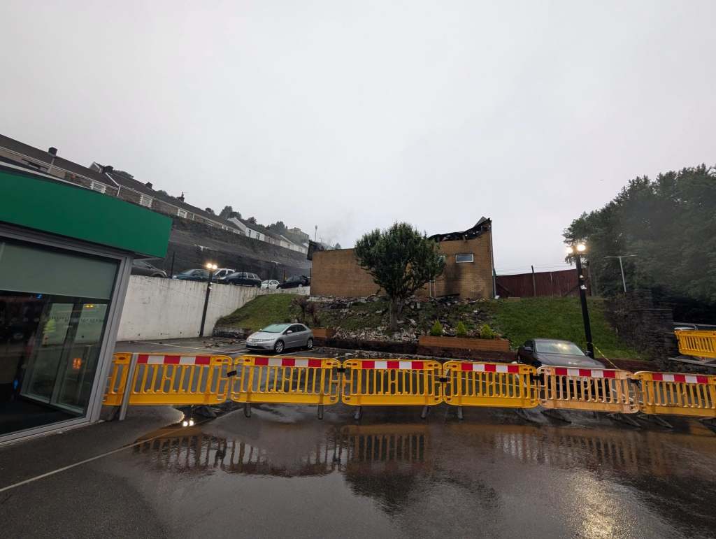 The fire-damaged industrial unit with its roof largely destroyed, behind yellow safety barriers next to the Enterprise Rent-A-Car premises