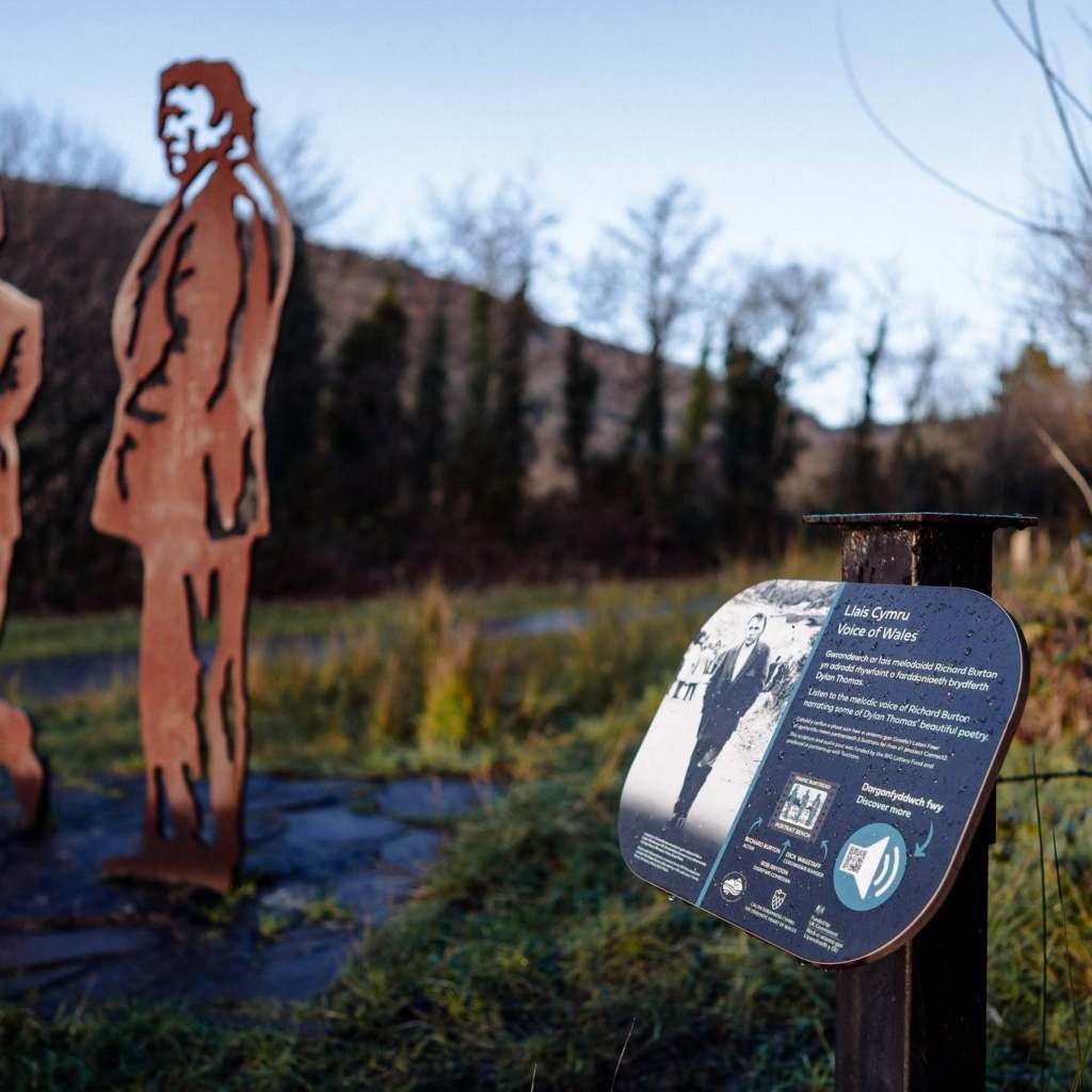 A rusted steel cut-out sculpture of Richard Burton beside a bilingual Voice of Wales information panel in the Afan Valley