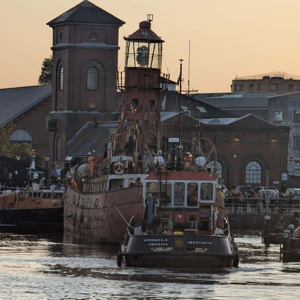 The Helwick lightship bow-on with her light tower rising in front of the Pump House, as the tug Veronica D guides her through the marina