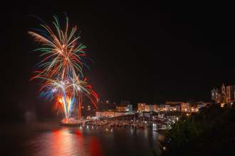 Fireworks exploding over Tenby Harbour at night during a Summer Spectacular event, with crowds lining the harbour walls and the town's pastel houses lit up behind moored boats