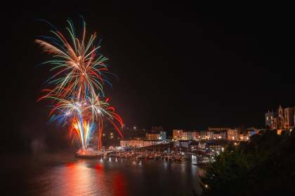 Fireworks exploding over Tenby Harbour at night during a Summer Spectacular event, with crowds lining the harbour walls and the town's pastel houses lit up behind moored boats