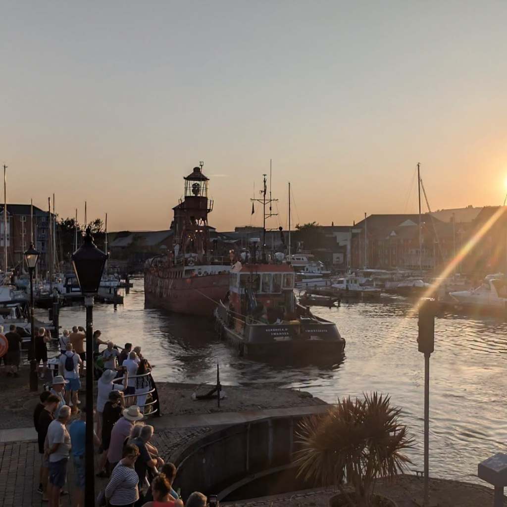 Crowds watching from the marina lock edge as the Helwick lightship is towed towards them at sunset