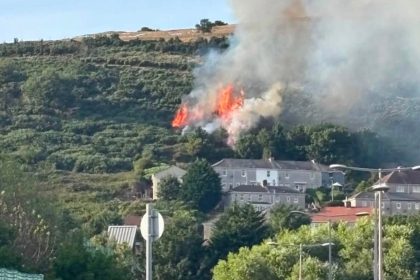 Flames and smoke on the green hillside of Kilvey Hill above rows of houses, with two onlookers watching from a street below