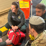 Three Welsh Ambulance Service staff in green uniforms and a soldier in camouflage and a beret kneel in a circle around a red medical response bag, with a clinical waste stand and a stretcher behind them.