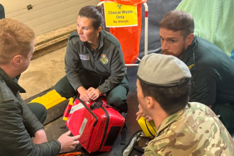Three Welsh Ambulance Service staff in green uniforms and a soldier in camouflage and a beret kneel in a circle around a red medical response bag, with a clinical waste stand and a stretcher behind them.