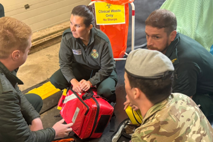 Three Welsh Ambulance Service staff in green uniforms and a soldier in camouflage and a beret kneel in a circle around a red medical response bag, with a clinical waste stand and a stretcher behind them.