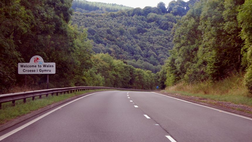 A white roadside sign reading Welcome to Wales, Croeso i Gymru, with a red dragon, beside a tree-lined dual carriageway