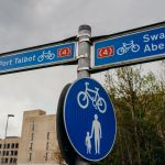 A blue cycle route signpost showing directions to Swansea and Port Talbot along National Cycle Network Route 4, with a shared-use path sign below.