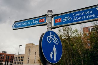 A blue cycle route signpost showing directions to Swansea and Port Talbot along National Cycle Network Route 4, with a shared-use path sign below.