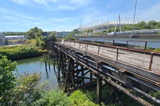 The weathered timber deck and trestles of the Morfa Bascule Bridge over the River Tawe, with the Swansea.com Stadium in the background